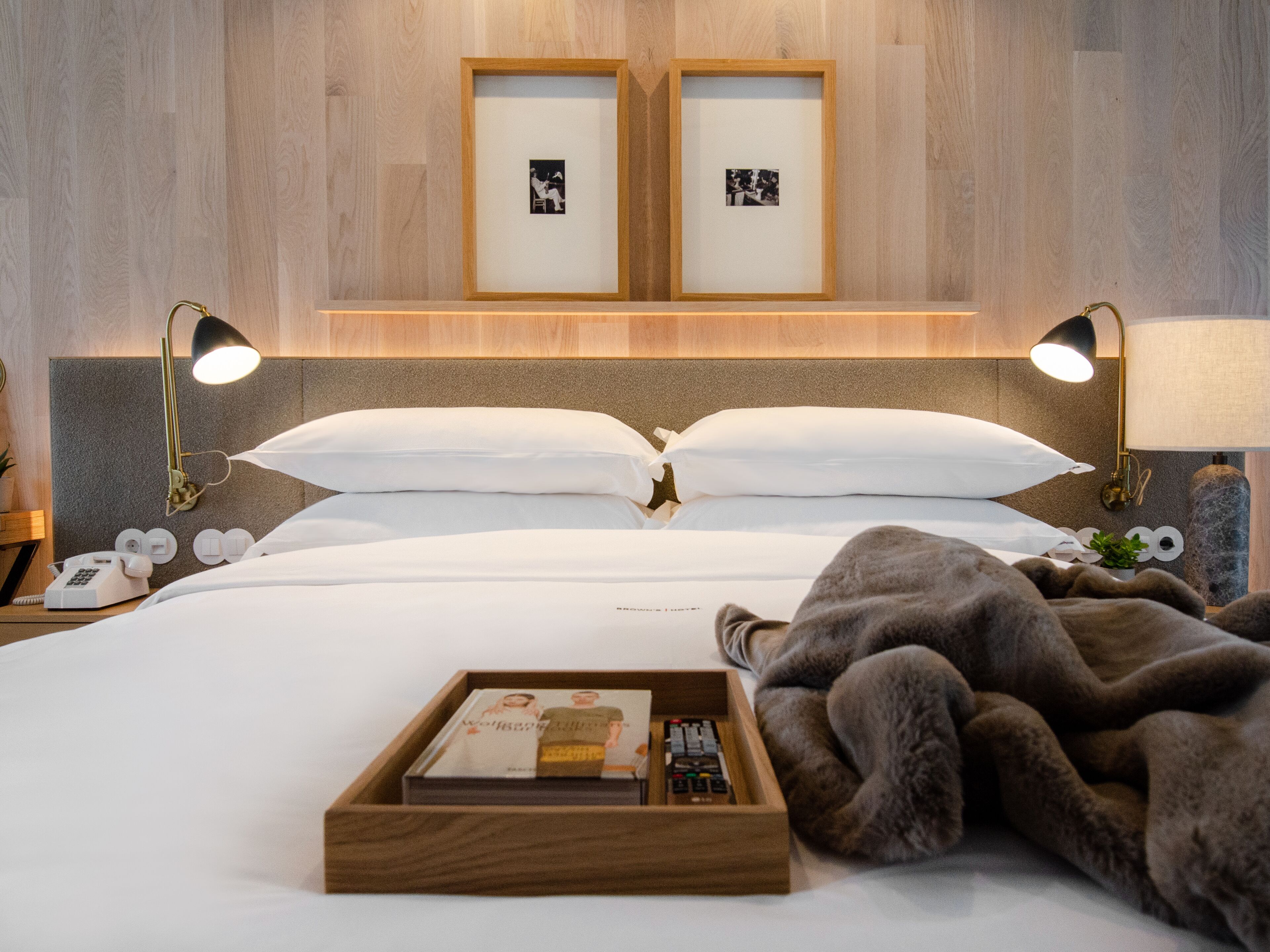 A neatly made bed with white linens, two framed pictures on the wall, bedside lamps, a bedside telephone, and a tray with books and a remote control.