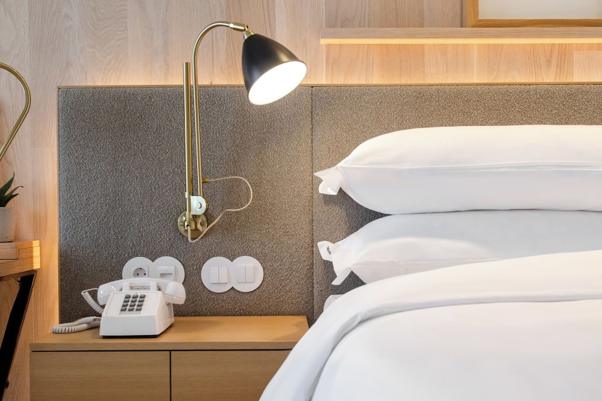 Ein Hotelzimmer mit einem Bett, einem Nachttisch mit Lampe, einem Telefon und einem an der Wand befestigten Lichtschalter.