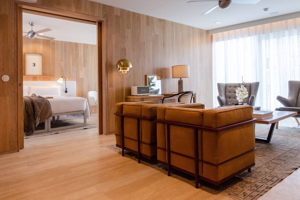 Ein modernes Hotelzimmer mit Holzboden, einem Bett, einer Sitzecke mit Sofa und Sesseln, einem Schreibtisch mit Lampe und einem Deckenventilator.