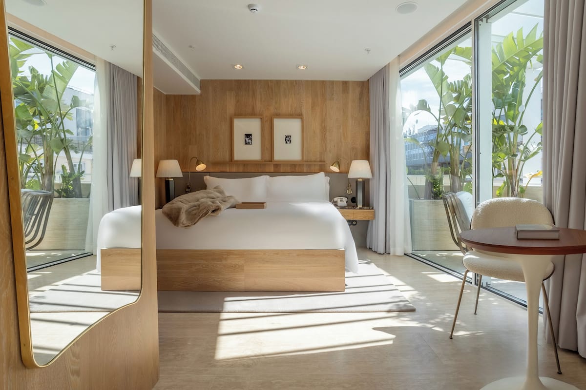 Ein modernes Hotelzimmer mit einem großen Bett, Nachttischen, einem Sessel und Blick auf Grün durch große Fenster.