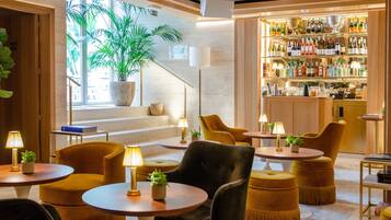 A modern bar area with a staircase leading to a balcony, furnished with round tables and chairs, and a variety of bottles and glasses on shelves.
