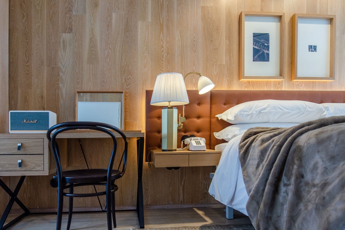 Ein Hotelzimmer mit einer Wandpaneele aus Holz, einem Bett mit weißer Bettwäsche, einem Nachttisch mit Lampe, einem Sessel, einem Schreibtisch mit Telefon und gerahmten Bildern an der Wand.