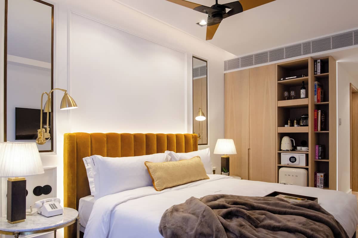 Ein Hotelzimmer mit Bett, Deckenventilator, Nachttisch mit Lampe, Telefon und Bücherregal.