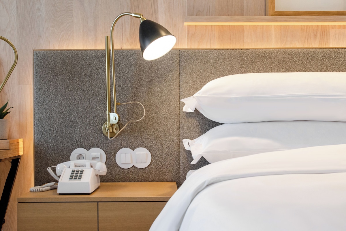 Une chambre d’hôtel avec un lit, une table de chevet avec une lampe, un téléphone et un interrupteur fixé au mur.