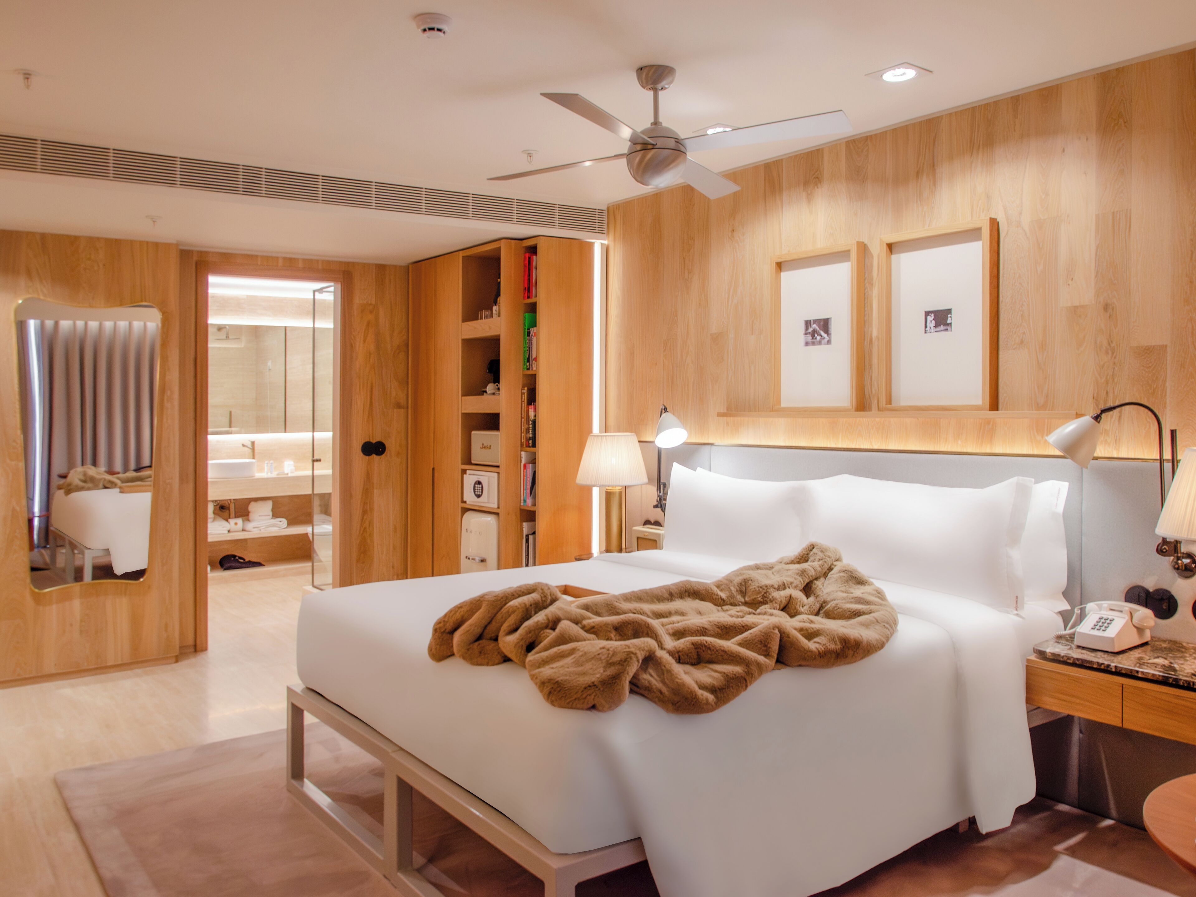 A modern hotel room with a large bed, wooden paneling, a ceiling fan, a bedside table with a lamp, and a nightstand with a phone.