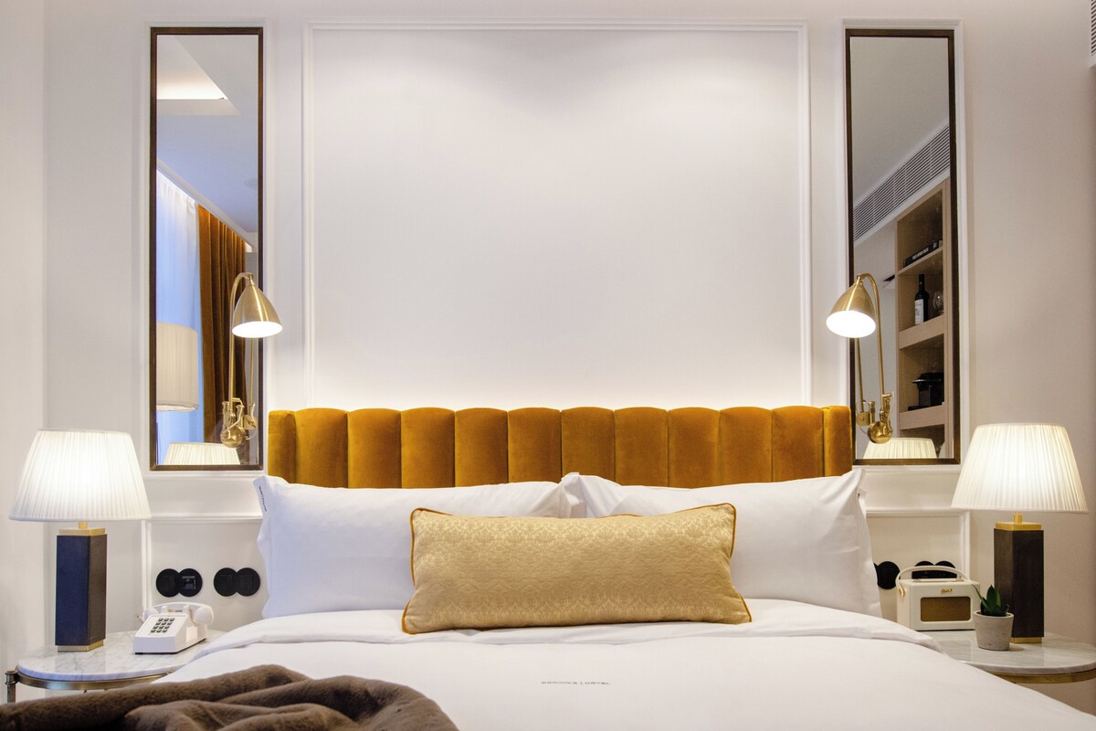 Une chambre d’hôtel comprenant un lit, deux lampes de chevet, un téléphone, un miroir et une table de chevet avec une lampe et une petite plante.