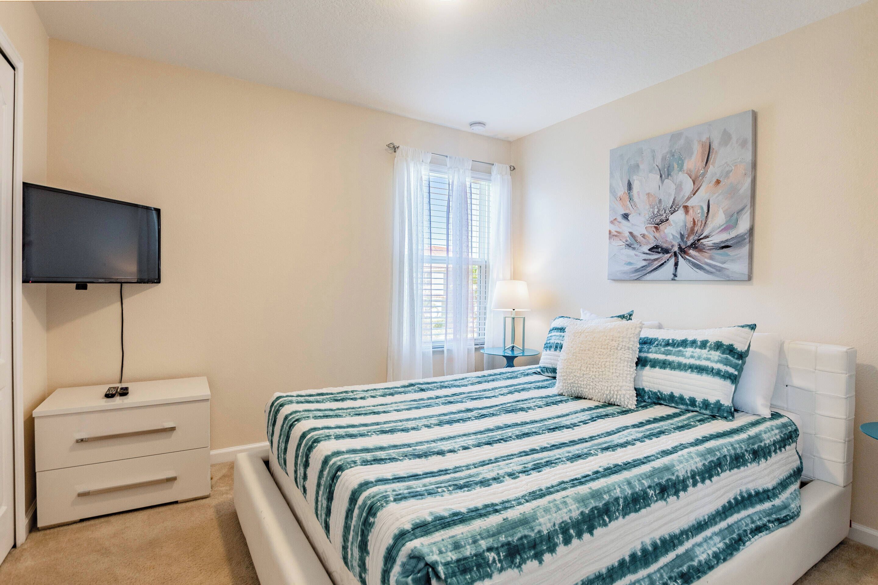Un dormitorio con cama, televisión, mesita de noche, lámpara y ventana con cortinas.
