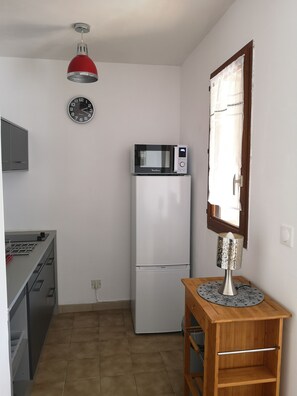 A compact kitchen with a microwave on top of a white refrigerator, a clock on the wall, and a small wooden table with a decorative item. - Charming Studio in South Bastia (Bastia)