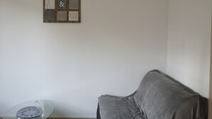 Una habitación con un sofá gris, una mesita redonda con superficie de vidrio y un tablón de anuncios en la pared.