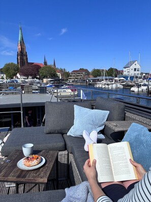 Eine Person entspannt auf einer Terrasse mit einem Buch und einem Teller Essen, mit Blick auf eine Marina mit Booten und einer Kirche im Hintergrund.
