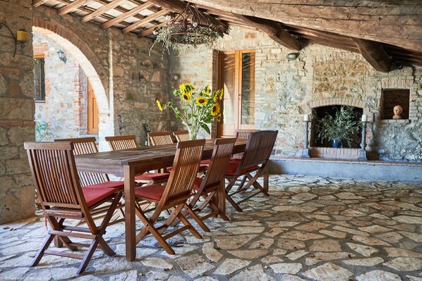 Un coin repas rustique, avec un sol en pierre, une table et des chaises en bois, un vase rempli de tournesols et une cheminée agrémentée d’une plante.