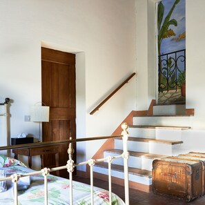 A bedroom with a wooden door, a staircase with a metal railing, a floral bedspread, and a suitcase on the floor.