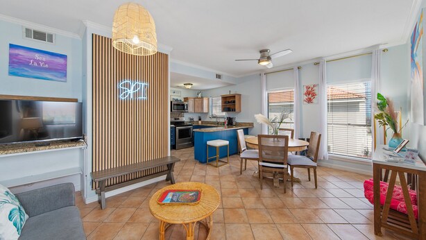 Una cocina moderna con una isla azul, un comedor con una mesa redonda y sillas, una sala de estar con un sofá y televisor, y un ventanal con persianas.
