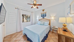 Un dormitorio con una cama, ventilador de techo, ventana con cortinas, mesita de noche con lámpara y un adorno en la pared.