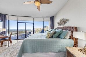 A bedroom with a large bed, a ceiling fan, a view of the ocean, a balcony with chairs, and a desk.