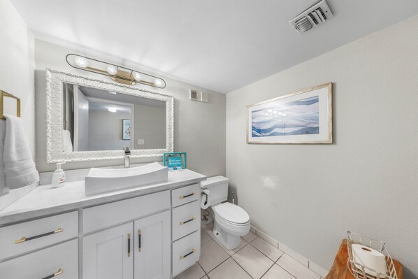 A modern bathroom with a white vanity, a large mirror, a framed picture on the wall, and a toilet.