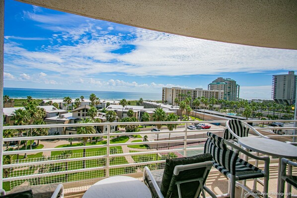 A balcony with a view of the ocean and a resort area, including palm trees and a parking lot.
