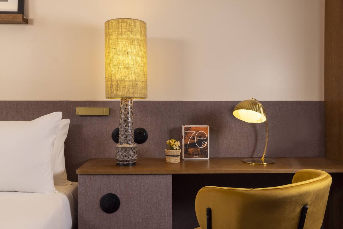 A hotel room with a bed, a desk with two lamps, a small vase with flowers, and a framed picture on the desk.