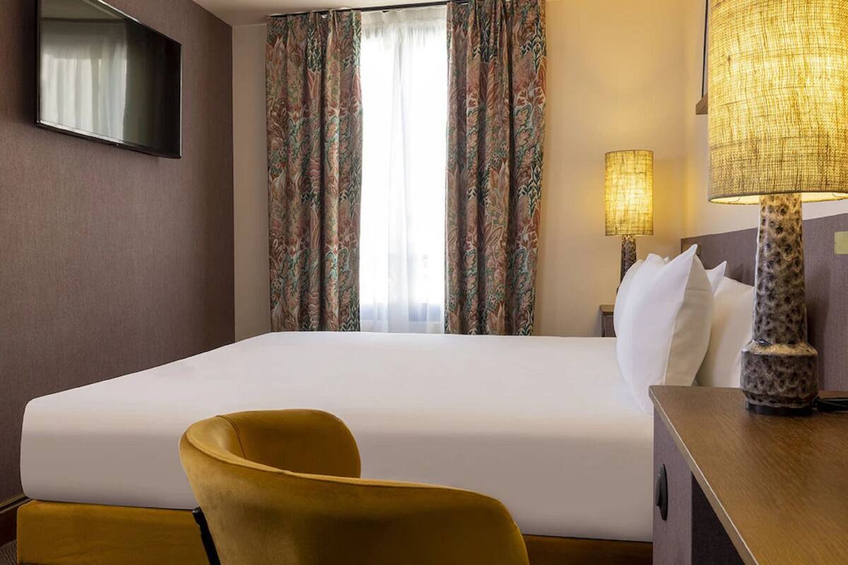A hotel room with a large bed, a television mounted on the wall, a desk with a lamp, and a chair.