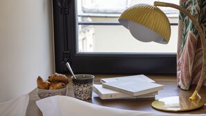 A table with croissants, a cup, books, and a lamp near a window with a view of buildings. - Hotel Ernest (Paris)