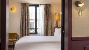 A hotel room with a large bed, a chair, a window with curtains, a lamp, and a door with a number. - Hotel Ernest (Paris)