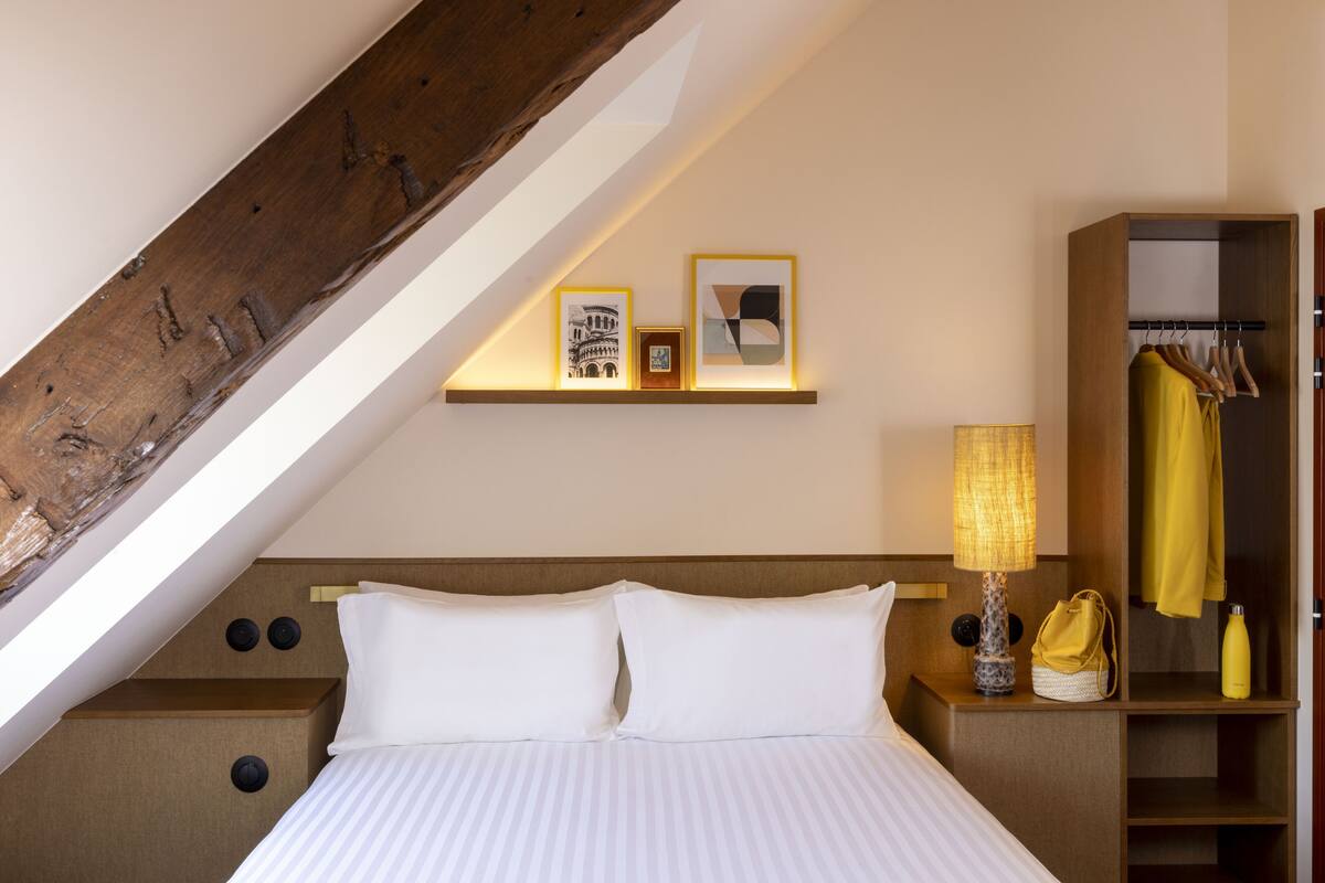 A hotel room with a bed, a nightstand with a lamp, a shelf with framed pictures, and a wardrobe with hanging clothes.