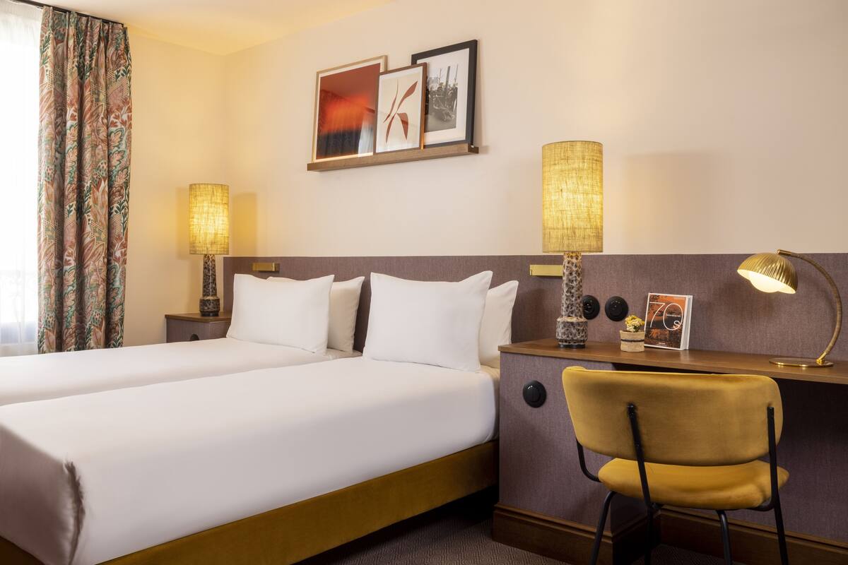 A hotel room with two beds, a desk, a chair, and two lamps.