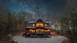 Eine zweistöckige Holzhütte mit Veranda, umgeben von Bäumen und einem sternenübersäten Nachthimmel.