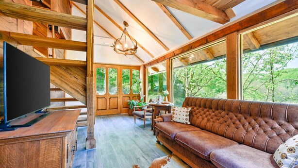 Un salon spacieux avec un grand canapé marron, des poutres en bois, un téléviseur et une vue sur la verdure environnante.