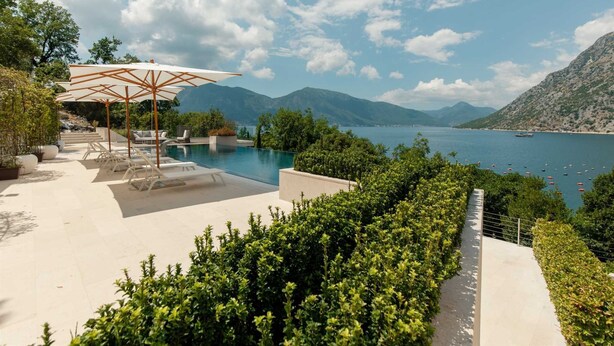 Un espace piscine agrémenté de chaises longues et de parasols, entouré d’une végétation luxuriante et offrant une vue sur un lac où naviguent des bateaux.