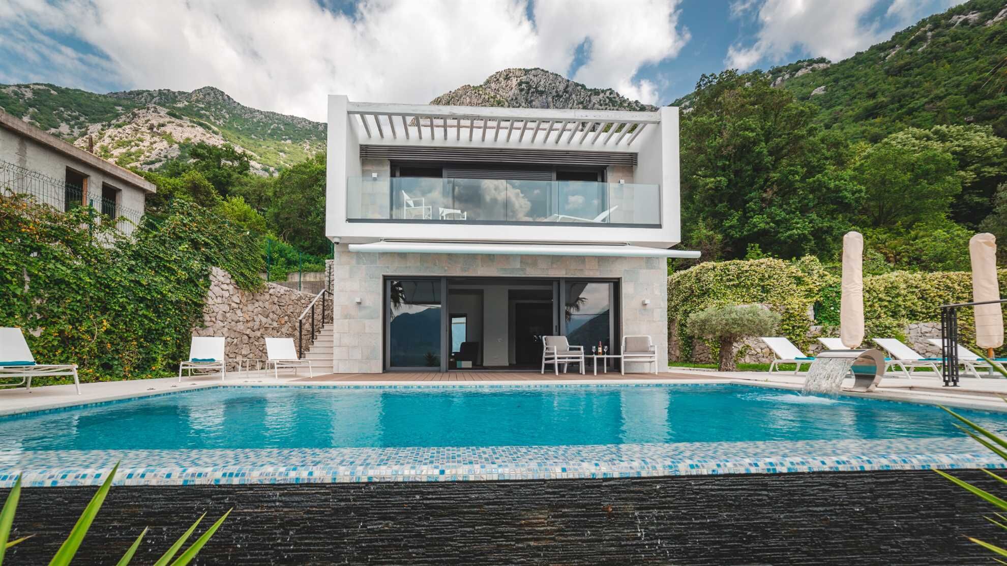 A modern two-story house with a swimming pool, surrounded by lush greenery and mountains.