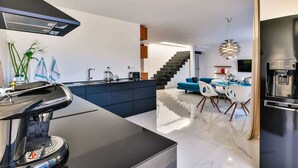 Une cuisine moderne équipée d’une machine à café, un coin repas avec un canapé bleu et des chaises blanches, et un escalier menant à un autre niveau.