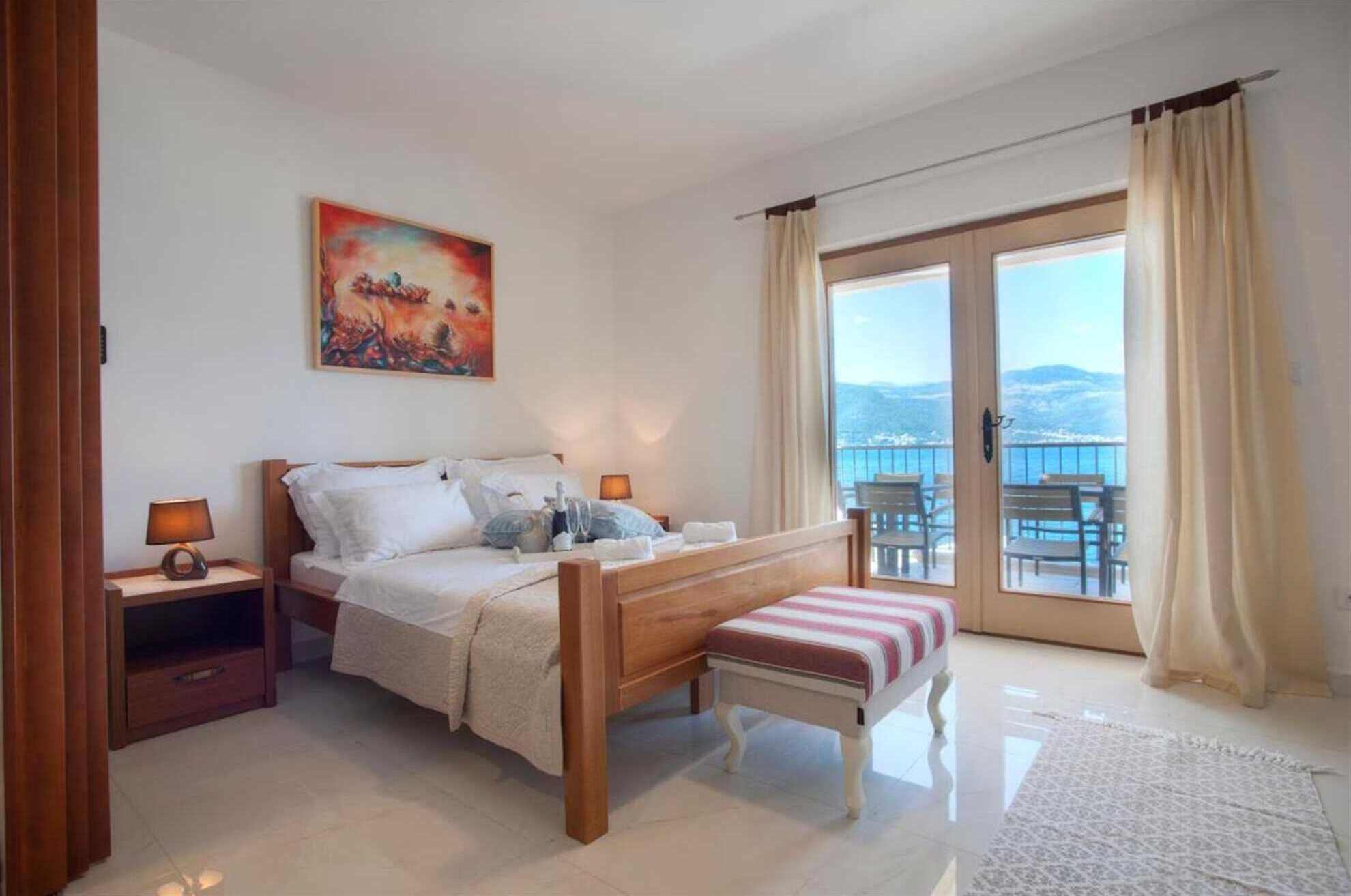 A bedroom with a bed, bedside tables, a bench, a painting, and a balcony with a view.
