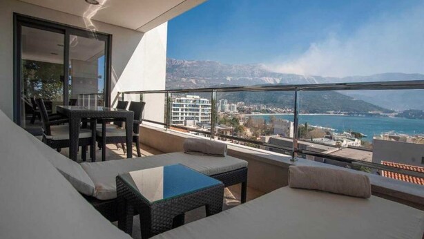 A balcony with a sofa, a table, and chairs, overlooking a coastal cityscape with mountains in the background. - Alexandar Suits & Spa 401 (Budva)