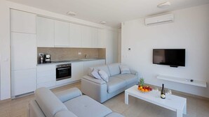 Ein zeitgemäßer Wohnraum mit Küche, einem grauen Sofa, einem Couchtisch mit Obst und Wein sowie einem an der Wand befestigten Fernseher.