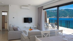 Ein modernes Hotelzimmer mit Balkon, Flachbildfernseher, Sofa, Esstisch und Blick auf Meer und Berge.