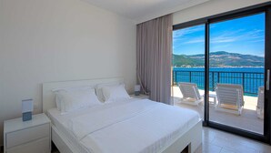 Ein Hotelzimmer mit Bett, Nachttisch, Lampe, einem großen Fenster mit Blick auf Meer und Hügel sowie einem Balkon mit Stühlen.