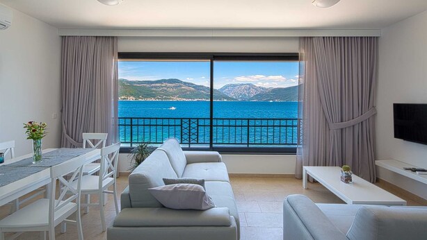 Ein modernes Wohnzimmer mit einer Couch, einem Esstisch und einem Fernseher. Ein großes Fenster bietet Blick auf das Meer und die Berge.