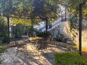 Un escalier en pierre mène à un bâtiment, qui est entouré d’une végétation luxuriante et doté d’un espace extérieur abrité avec des chaises et une table en métal.
