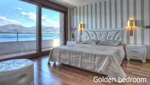 Een slaapkamer met een groot raam, een bed met een witte kap, twee nachtkastjes met lampen en uitzicht op zee.