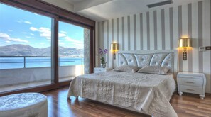 Une chambre avec une grande fenêtre, un lit doté d’une tête de lit blanche, deux tables de chevet avec des lampes et une vue sur la mer.