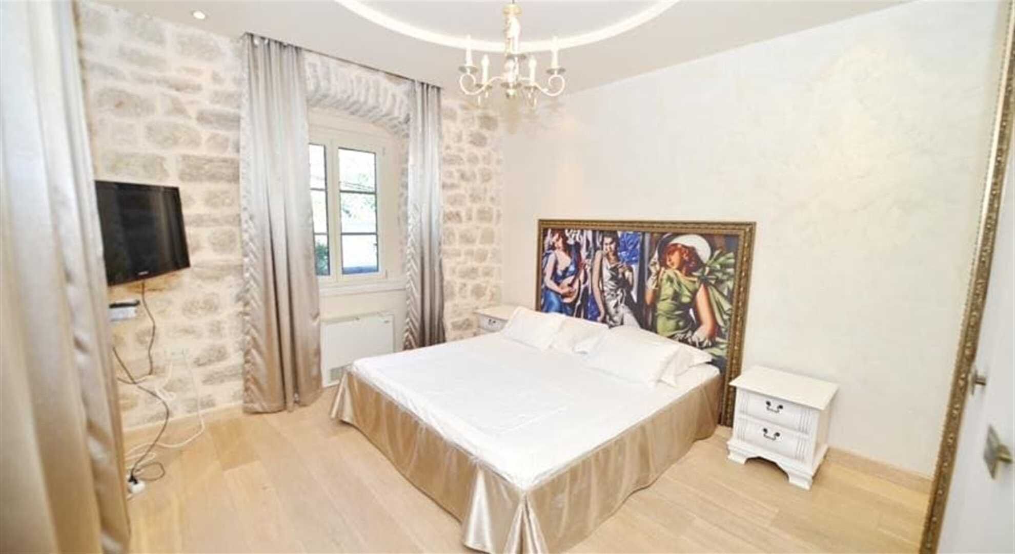 A bedroom with a large bed, a window with curtains, a painting on the wall, and a chandelier.
