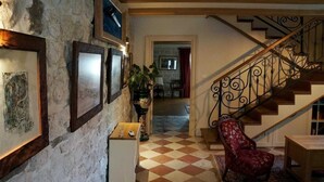 Un pasillo con piso de tablones de ajedrez, una escalera con barandales de metal ornamentado y cuadros en las paredes.