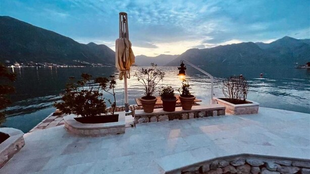 Una terraza con plantas en macetas, una mesa y vistas a un lago con montañas al fondo.