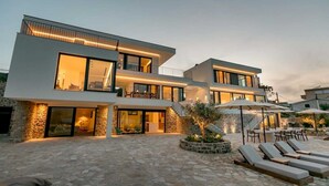Una casa moderna d'àmbit elevat amb façana de pedra, grans finestres i un pati amb gandules i ombrel·les.