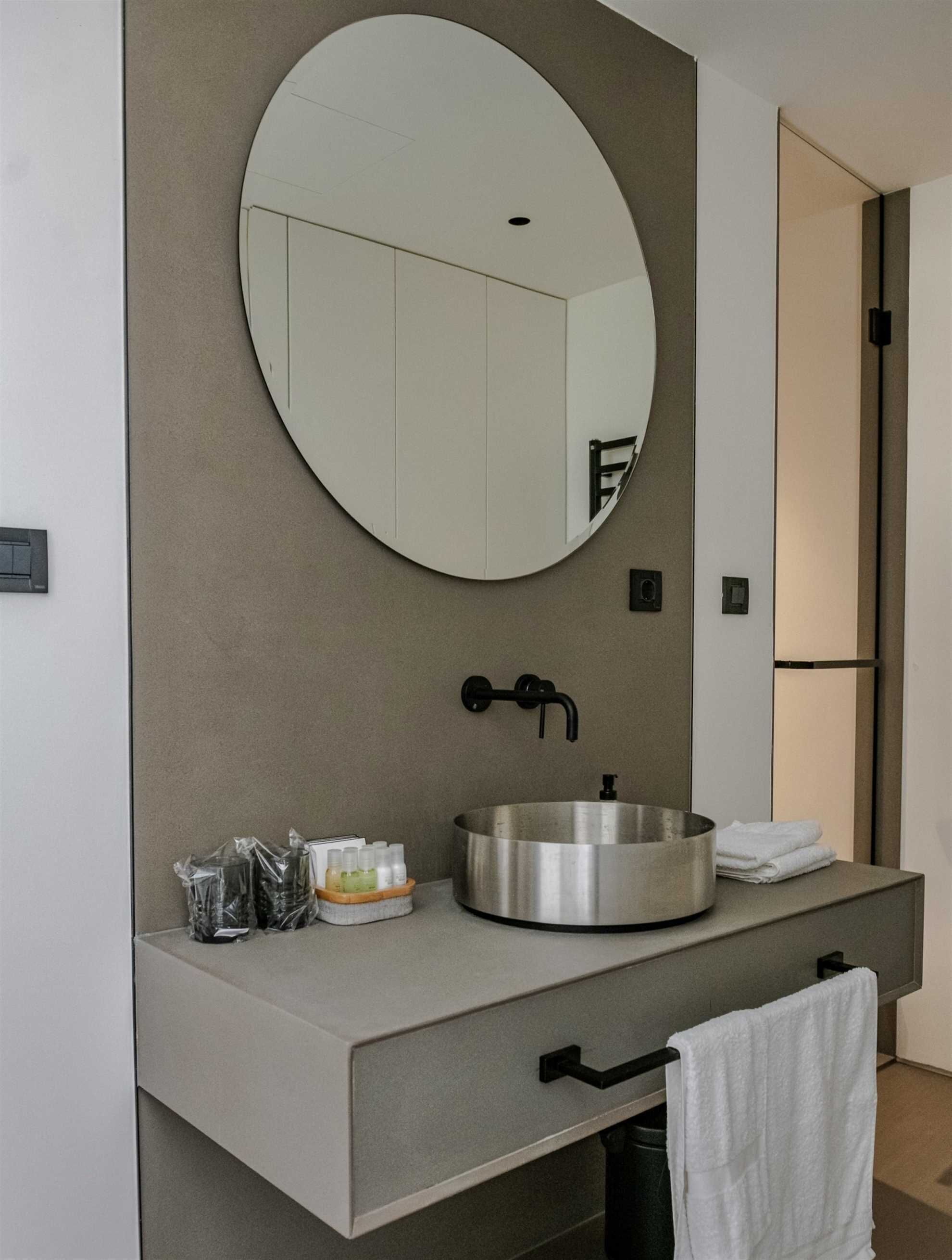 Un baño moderno con un espejo redondo, un lavamanos de acero inoxidable y un perchero para toallas.