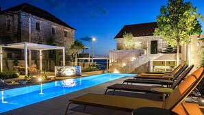 Una piscina con lettini, una zona relax coperta e edifici in pietra sullo sfondo.