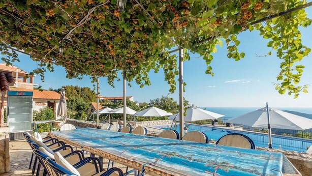 Ein Poolbereich mit langem Tisch und Stühlen, Sonnenschirmen und Blick aufs Meer.