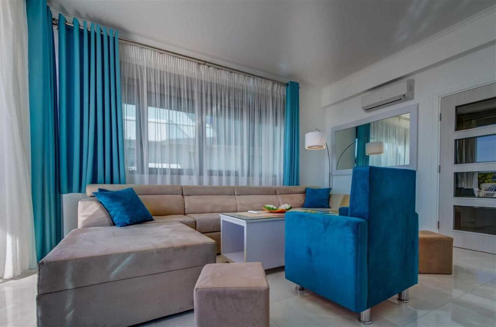 A modern living room with a beige sectional sofa, a blue armchair, a small white side table, and a large window with sheer curtains.