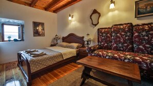 A room with a wooden ceiling, a bed with a wooden headboard, a floral sofa, a wooden table, and a window with a view. - Royal Villa Ana - 2 Bedroom Villa (Cetinje)